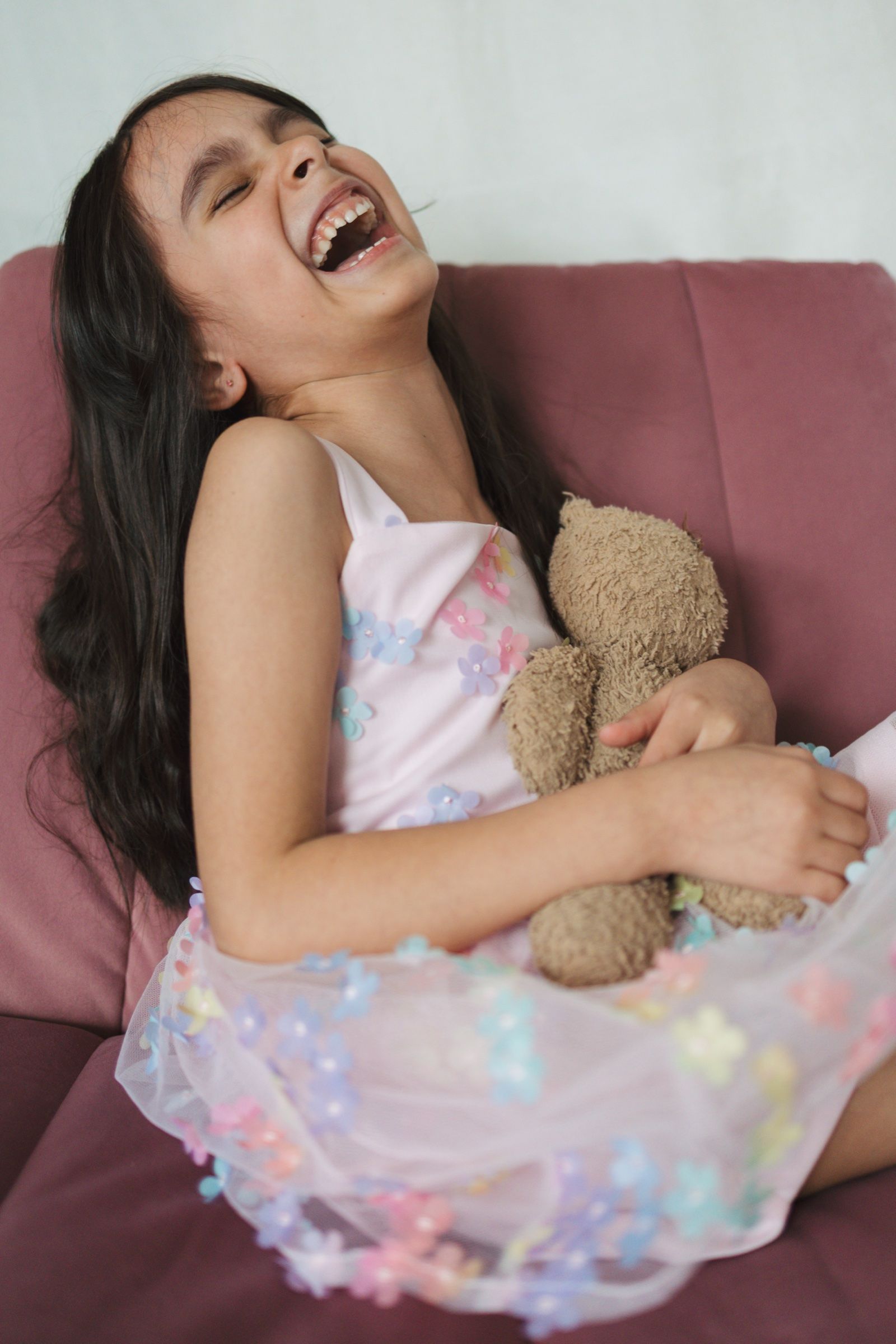 Young girl laughing while holding a teddy bear on a pink chair